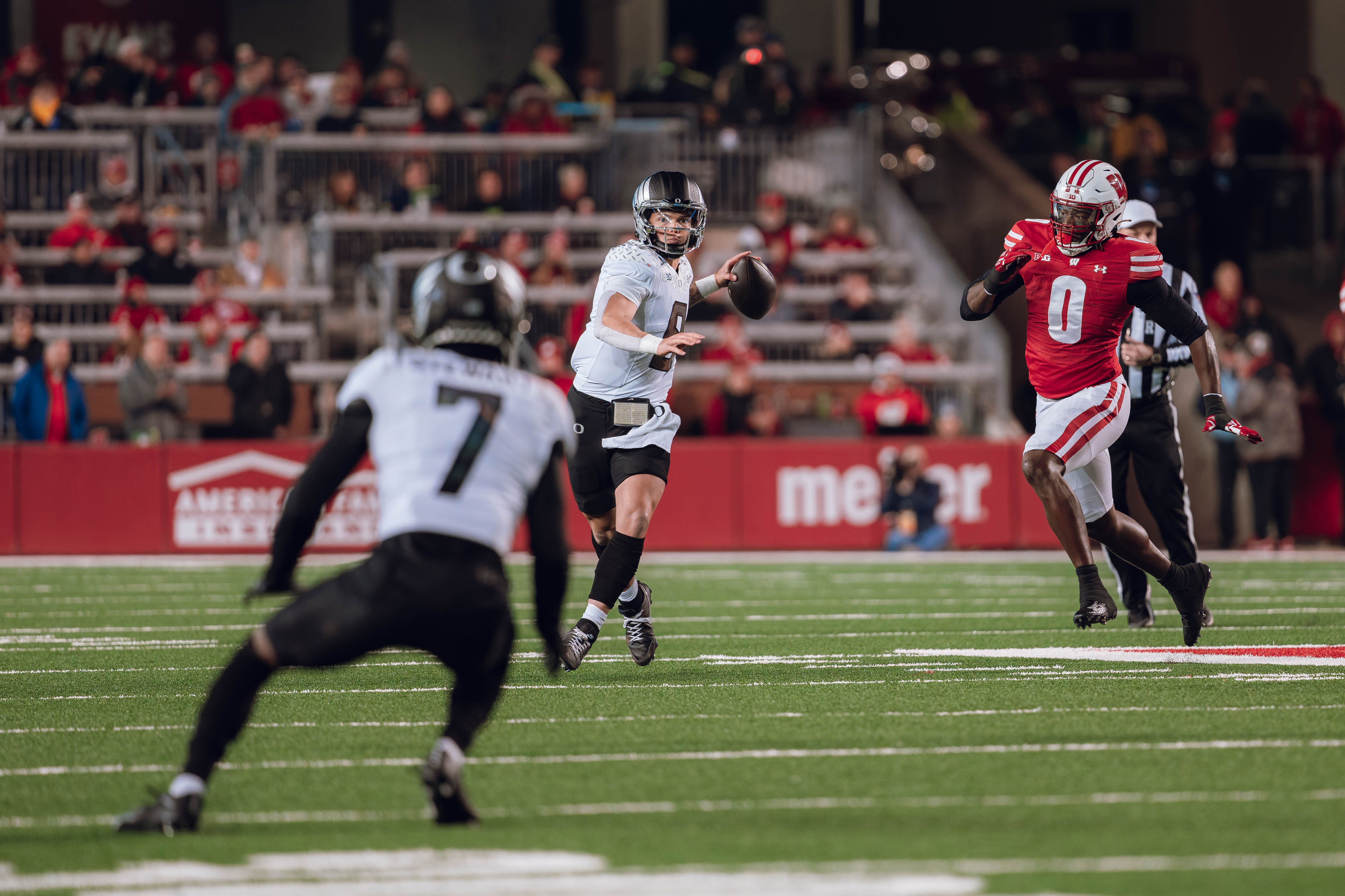 Oregon quarterback Dillon Gabriel #8 looks down field to Oregon wide receiver Evan Stewart #7 while being pursued by Wisconsin Badgers outside linebacker John Pius #0 at Camp Randall Stadium on November 16, 2024 in Madison, Wisconsin. Photography by Ross Harried for Second Crop Sports.