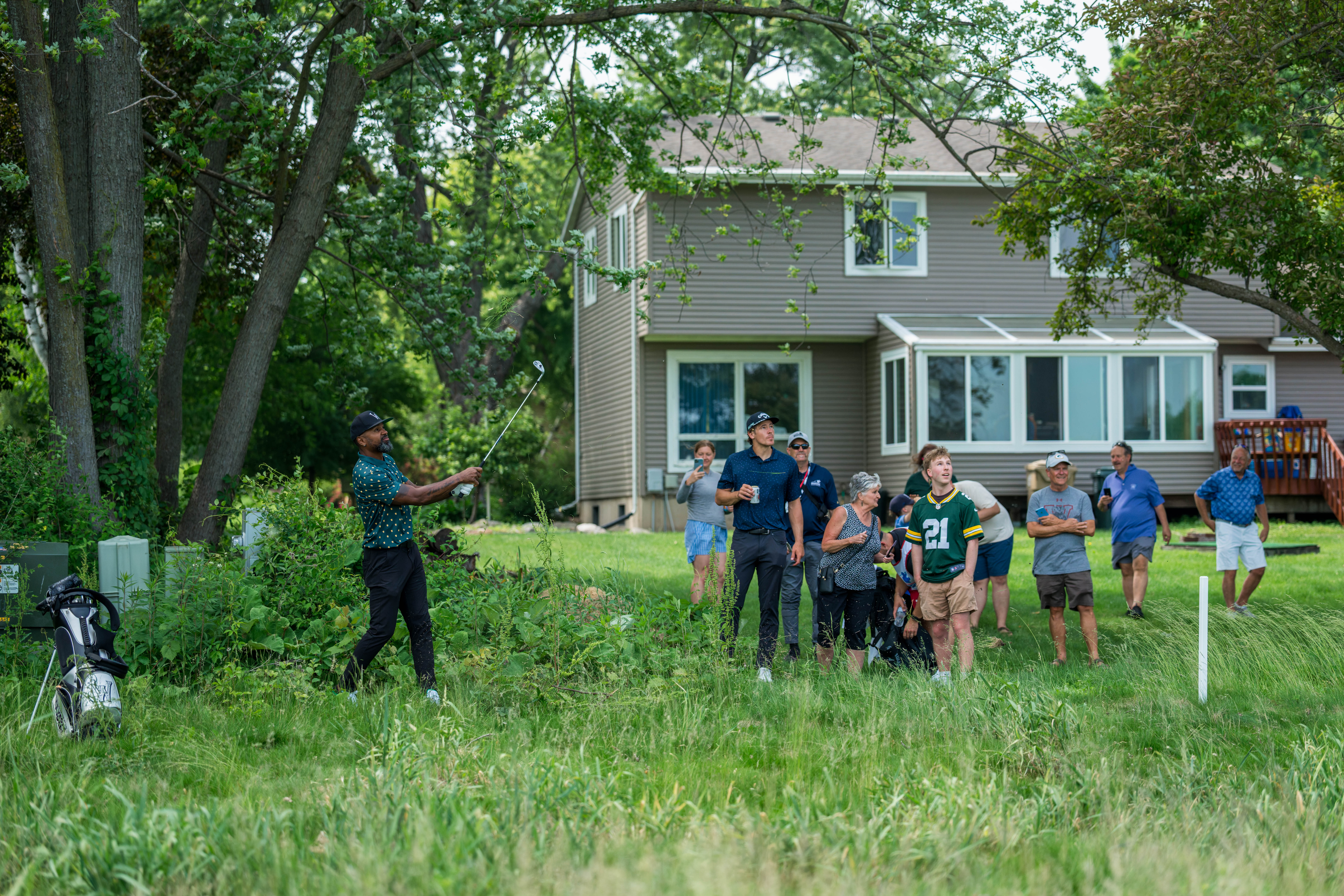 Charles Woodson (left) hits his second shot on hole 12 while Justin Herbert (center) mingles with a backyard party during round 2 of The American Family Insurance Championship at TPC Wisconsin in Madison, Wisconsin on June 7th, 2025. Photography by Ross Harried for Second Crop Sports.
